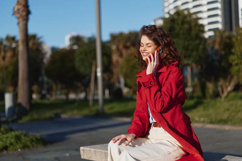 Young woman with curly hair wearing a red coat sits on a bench outdoors while talking on a mobile phone and smiling in the sunny urban park.