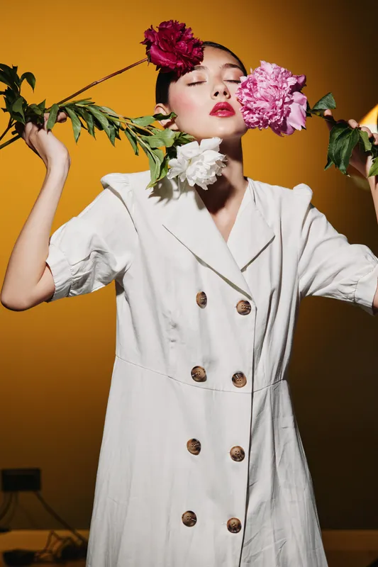 Female fashion model in a white linen dress holds colorful peonies near her face against a warm orange background. Creative beauty and floral concept for fashion editorial.