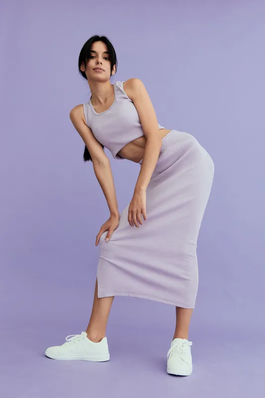 Young woman posing in a lilac knitted dress and white sneakers against a soft purple background, showcasing casual fashion with a relaxed and stylish vibe.