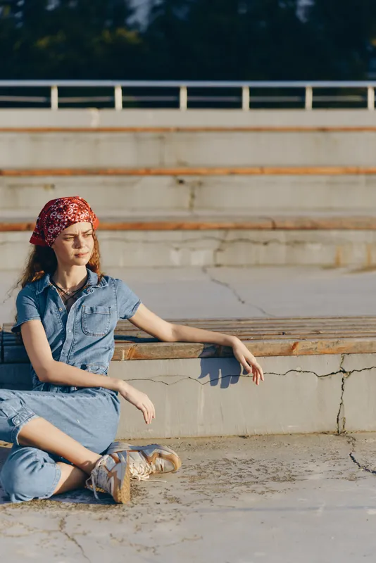 Young woman with red bandana sitting outdoors on concrete steps in casual denim outfit enjoying sunlight in a relaxed and peaceful mood.