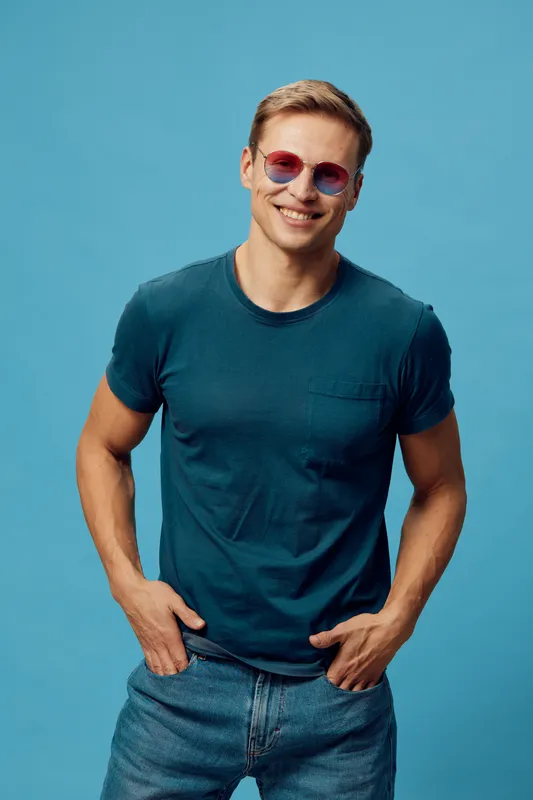 Smiling man with sunglasses wearing casual blue t-shirt and jeans looking cheerful against solid blue background in studio portrait. People lifestyle concept