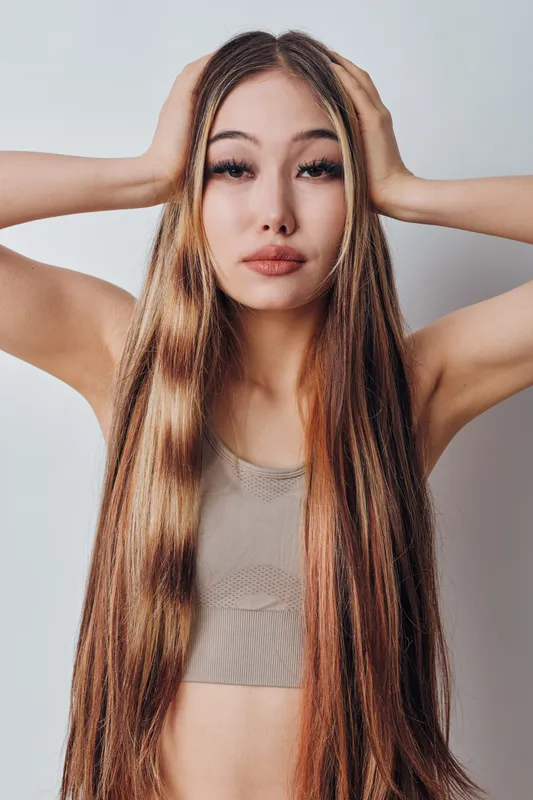 Woman with long straight hair posing with hands on head looking at camera wearing beige top in studio light on white background. Fashion, beauty and portrait closeup.