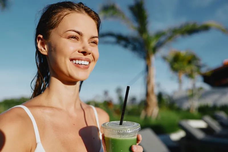 Happy woman enjoys a refreshing green smoothie outdoors, smiles warmly, with palm trees in the background, bright daylight, casual summer lifestyle moment by the poolside at a resort.