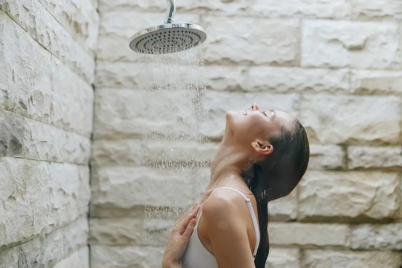 Woman enjoying outdoor shower under rainfall with stone wall background, wet hair, relaxed posture, and natural light creating calm spa atmosphere.
