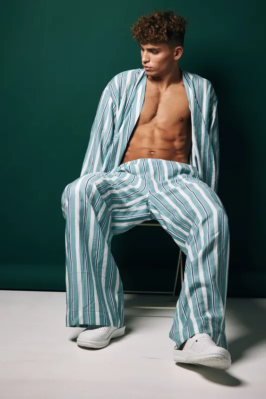 Young man sitting on chair in casual striped pajamas with open shirt posing with serious face on dark green background during indoor fashion photo session in studio.