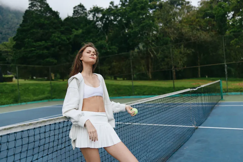 Young female tennis player in a sporty outfit, enjoying a moment of triumph on a tennis court surrounded by lush greenery, showcasing athleticism and confidence