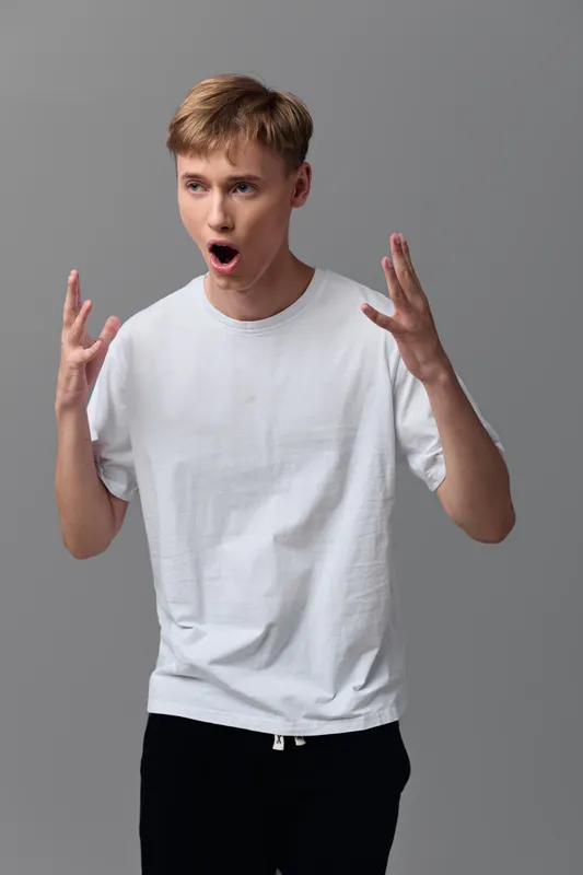 Young man in a white t shirt shows surprise with raised hands and open mouth against a gray studio backdrop, capturing a bold expressive moment for reaction and astonishment in advertising