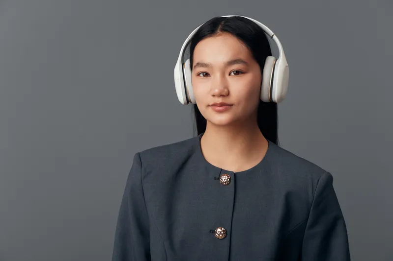 Young woman with headphones and long black hair standing against gray background wearing dark blazer, looking at camera with slight smile and professional appearance.