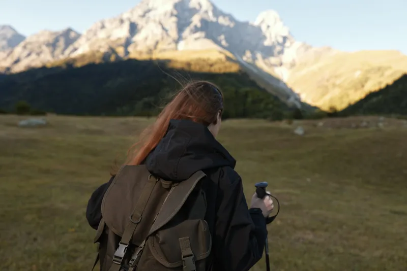 A lone traveler with a large backpack stands in a wide meadow as rugged mountains rise behind, creating a serene outdoor scene and a sense of exploration.