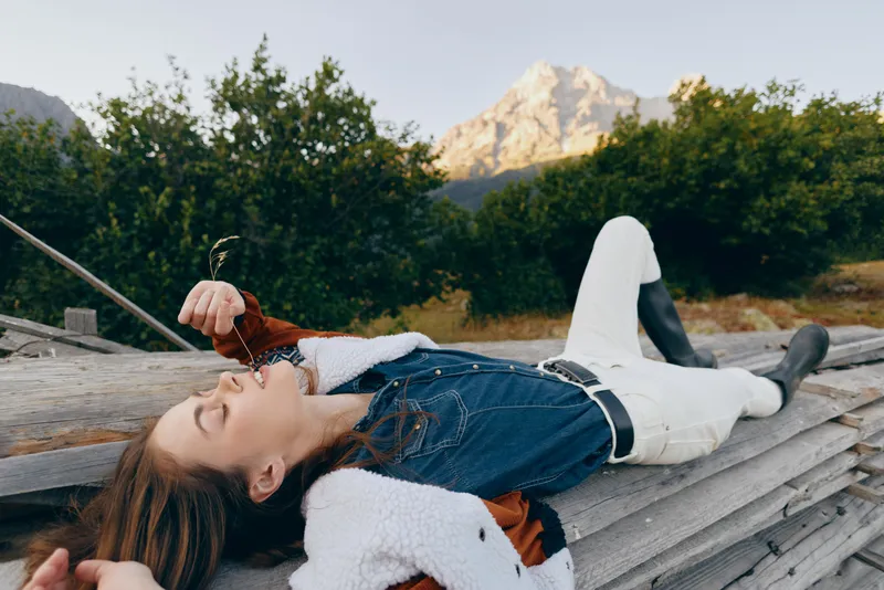 Woman lying on wooden logs eating wild berries in nature by mountain, wearing denim jacket and white pants, relaxed outdoors scene with boots and forest background.