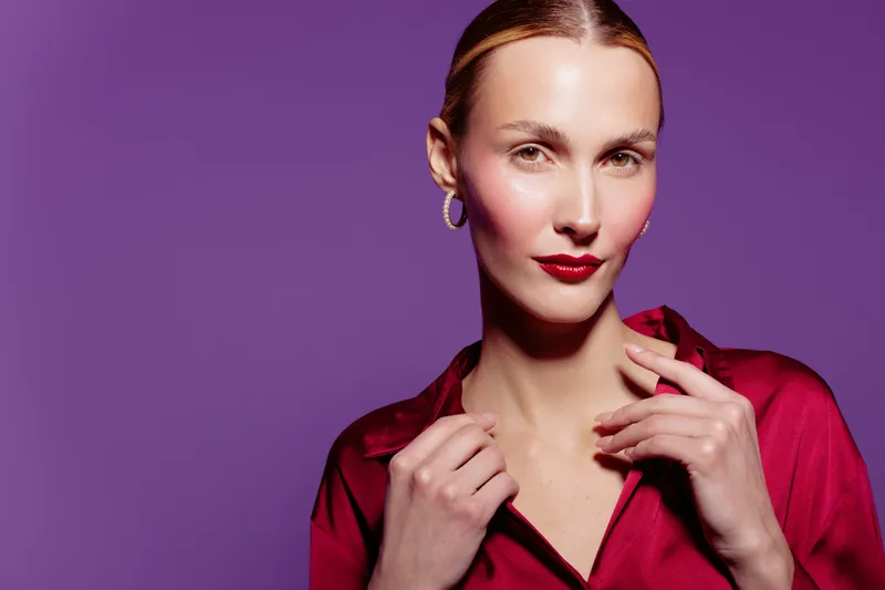 Confident woman with red lipstick and hoop earrings poses against purple background wearing red blouse. Studio portrait with soft lighting highlights facial features and elegant style.