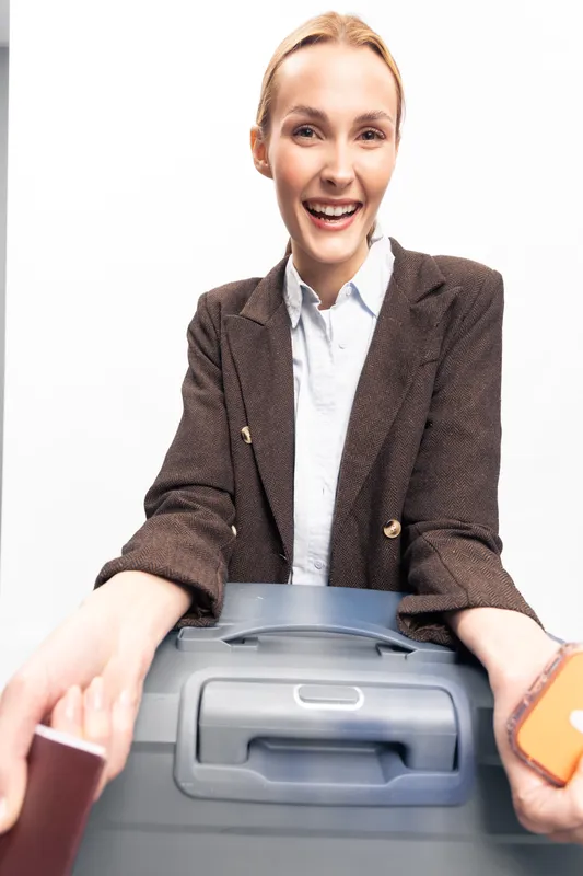 Smiling young woman with suitcase and passport ready for travel. She wears a brown blazer and holds a phone, expressing excitement and anticipation for a trip.