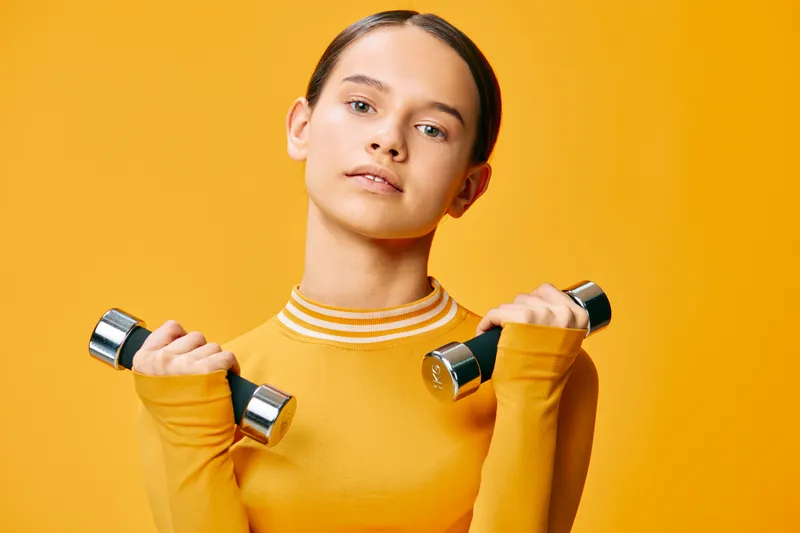 Young woman fitness portrait lifting chrome dumbbells in a yellow long sleeve top against a bright orange studio background, confident athletic pose and healthy lifestyle concept