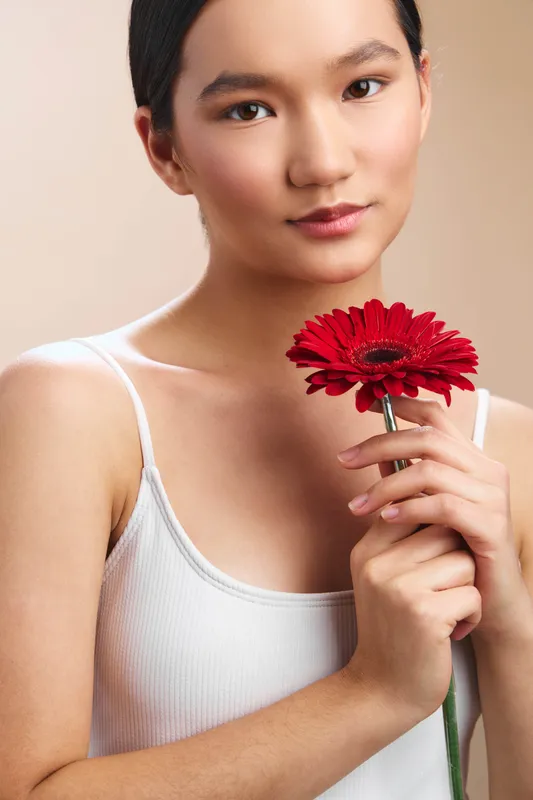 Asian woman holds a bright red gerbera flower close to her chest, wearing a white tank top. soft lighting and a neutral background create a calm, elegant portrait.