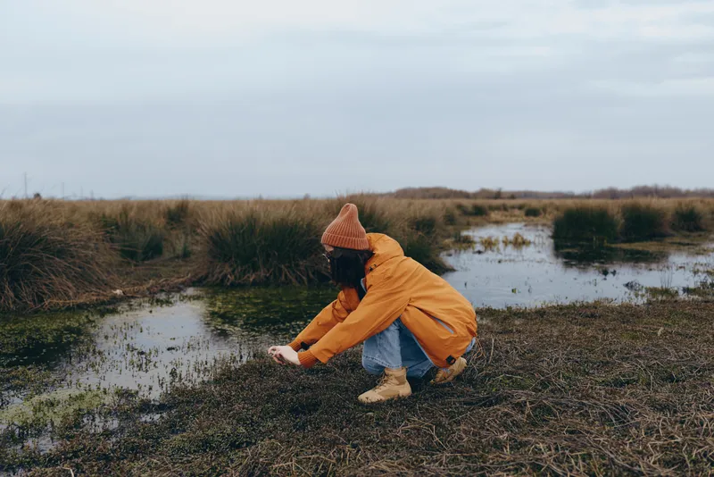Woman outdoors lifestyle kneels on marshy riverbank under cloudy sky, touching wet grass, nature walk concept for mindfulness and calm, orange jacket