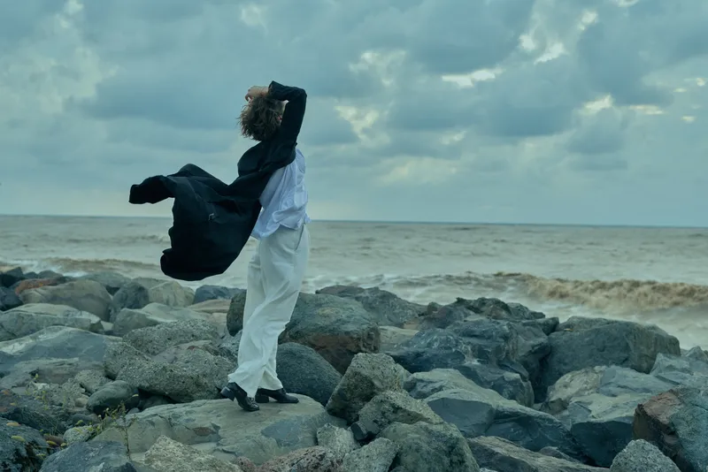 Wind sweeps a windswept figure along rugged rocks by the sea, white attire fluttering, dramatic ocean backdrop, moody sky, and scattered stones, capturing motion, solitude, and bold fashion in a