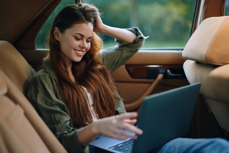 Young woman working on a laptop in a vintage car, showcasing joy and creativity while enjoying a relaxed outdoor setting with natural light.