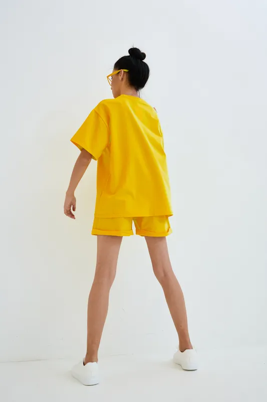Woman in yellow oversized outfit and shorts stands against white background with sporty white shoes and sunglasses, showcasing casual summer fashion style.