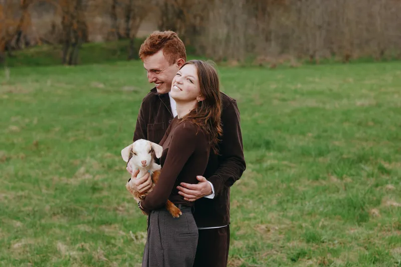 Couple outdoors with a goat kid in a field, embracing in a grassy park as sunlight warms their smiles, highlighting love, companionship, and a relaxed countryside lifestyle.