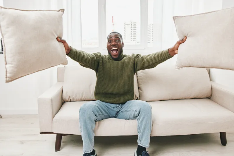 Joyful man celebrating at home with cushions in hand, wearing a green sweater and jeans, bright indoor setting evoking happiness and comfort