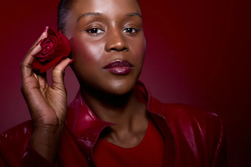 Close-up of a confident black woman with dark skin holding a red rose near her face. She wears a red leather jacket and lipstick, posing against a red background. Concept of beauty and elegance.