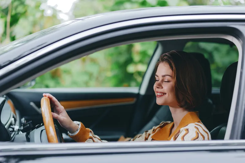 A cheerful woman drives a car, smiling with confidence as she rests her hand on the steering wheel and prepares for a journey.
