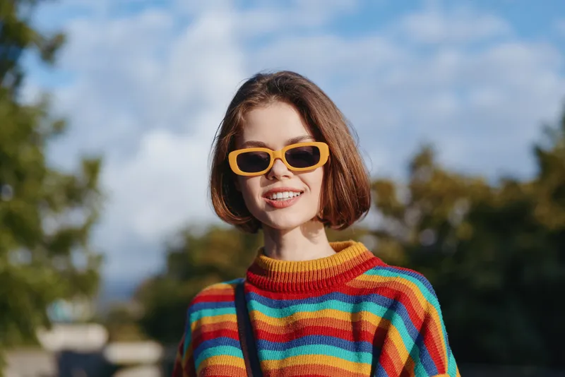 Woman in her mid-20s smiling in a colorful striped sweater and yellow sunglasses, short brown hair, casual outfit, outdoor portrait in a sunny park with lifestyle travel vibe.