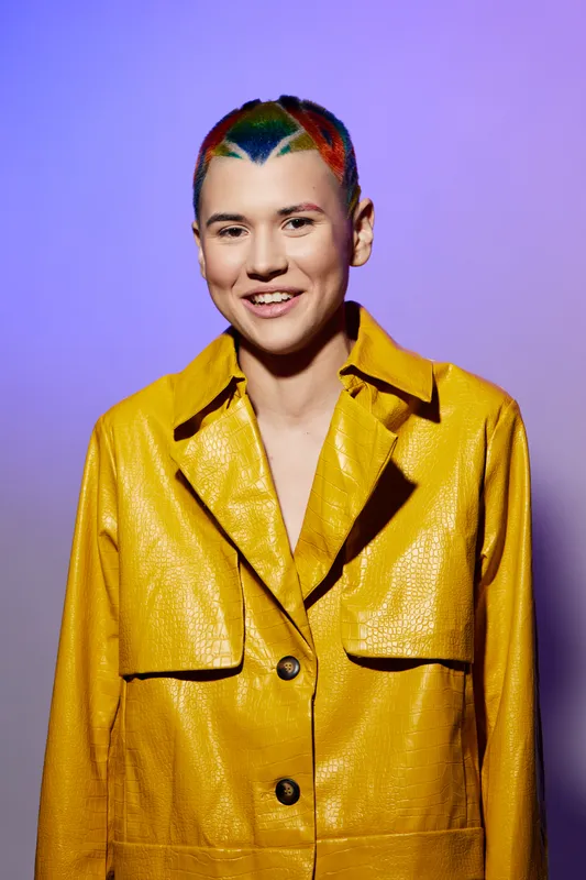 Young woman with colorful short hair wearing a shiny yellow jacket, smiling confidently against a vibrant purple background, showcasing bold fashion and unique style.