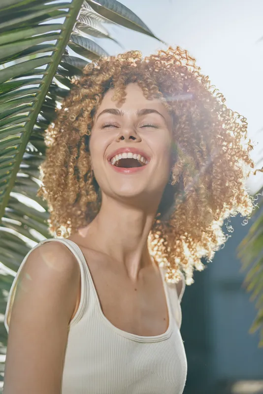 Smiling woman with curly hair enjoys sunny day outdoors near tropical palm leaves, wearing white top, radiating happiness and warmth with natural light.