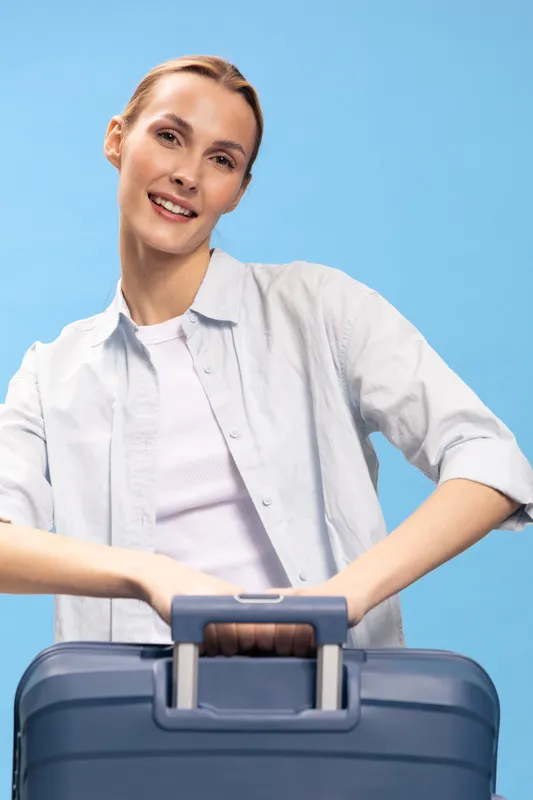 Smiling woman in casual white shirt holding blue suitcase handle against blue background. Travel concept with relaxed mood and positive female traveler ready for journey.