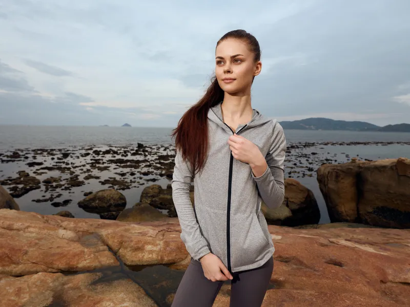 Young woman in sportswear standing on rocky shore, looking confident in activewear, enjoying nature and fitness, perfect for health and wellness themes