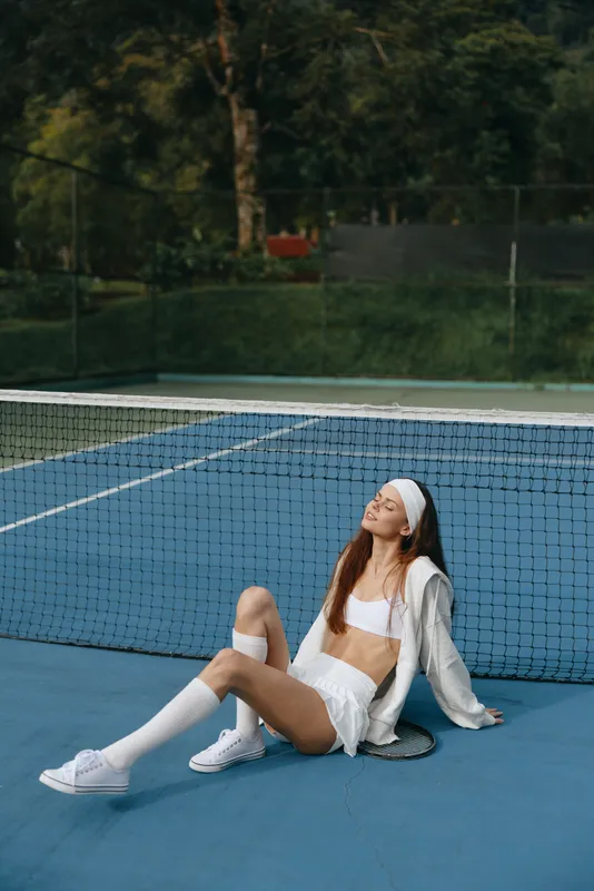 Young athletic woman in white sportswear relaxing on a blue tennis court with a joyful expression, embodying fitness and summer energy