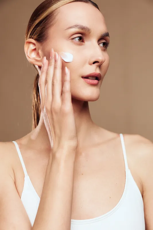 A young woman with clean skin applies white cream to her cheek with fingers. She wears a white tank top and looks away against a neutral background. Concept for skincare routine.