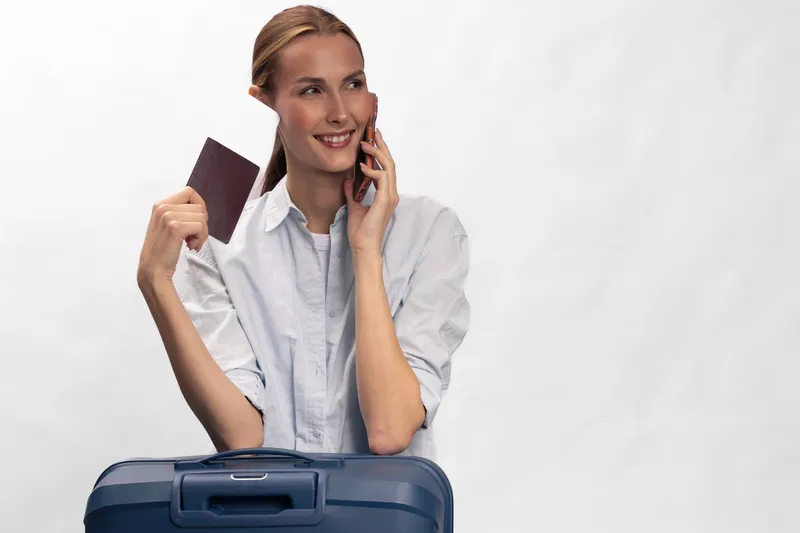 Happy woman with suitcase holds passport and talks on smartphone, ready for travel. Concept of tourism, vacation planning, and business trip preparation with positive mood and casual outfit.