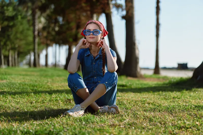 Lifestyle woman in modern boho-western denim street style sitting on grass with sunglasses and headscarf, relaxed outdoors under natural sunlight, film color effect.