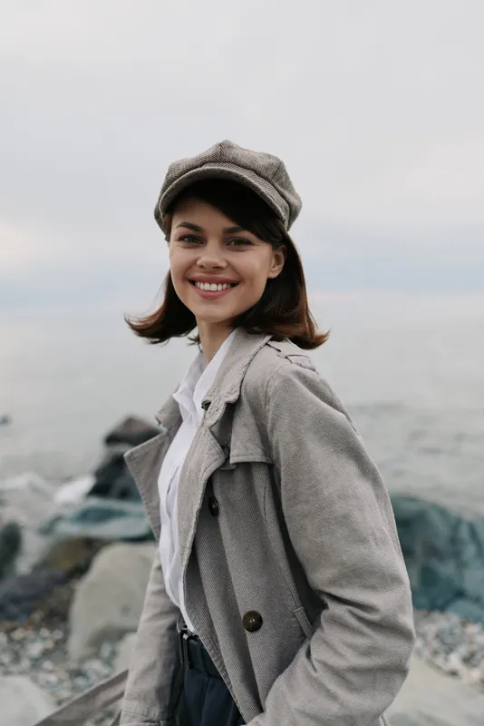 Young woman in grey coat and hat smiles warmly standing near rocky shore. Overcast sky and sea create calm atmosphere. Concept of autumn fashion and outdoor lifestyle.