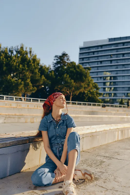 Lifestyle woman wearing modern boho-western denim street style sits outdoors relaxing in warm film color tones under clear sky in urban setting.
