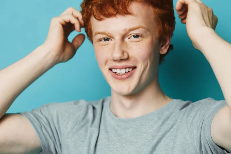 Young man with red hair and freckles smiling and wearing a gray t shirt on blue background in closeup portrait with natural expression and casual pose.