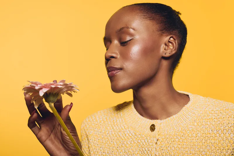 Dark skinned African woman in yellow sweater holds light pink flower close to face with closed eyes. Studio portrait on yellow background. Concept of natural beauty, calmness, and floral scent.