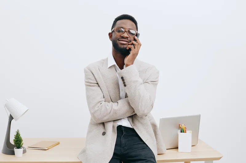 Professional young black man in smart casual attire leaning on desk with arms crossed and smiling at camera. Office background with laptop, stationery, and plant, emphasizing a cheerful, confident, mo