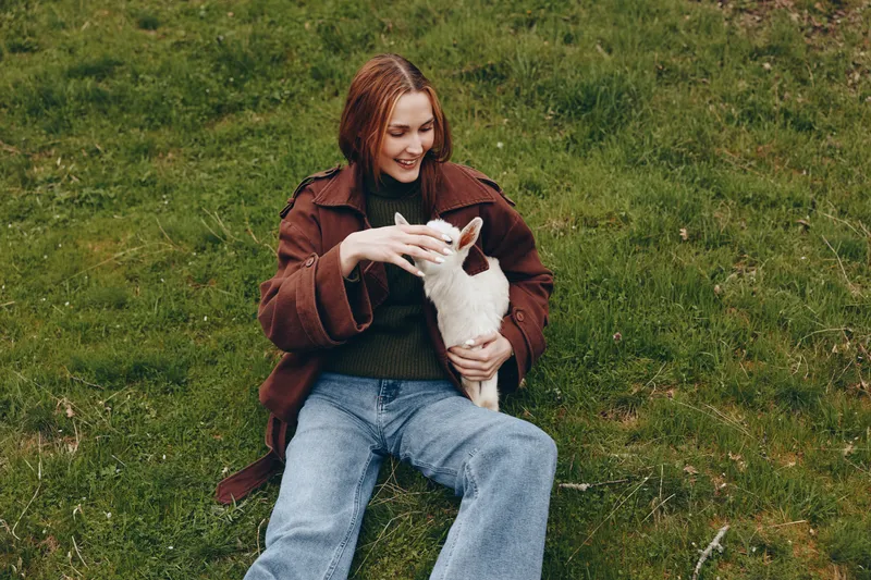 woman outdoors, goat kid, affectionate moment in nature, casual lifestyle vibe on grass, sunlight, relaxed posture, cozy coat, sweater, jeans, cheerful connection