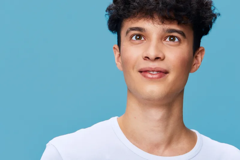 young man smiling with curly hair wearing white shirt looking up against blue background, positive expression and casual attire