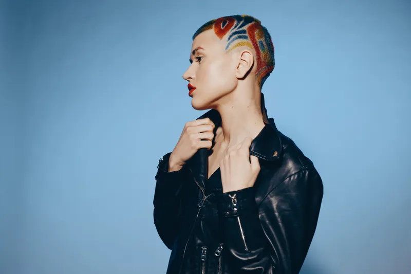 Stylish woman with colorful shaved hair and tattoo, wearing a black leather jacket, posing confidently against a soft blue background in a modern portrait.