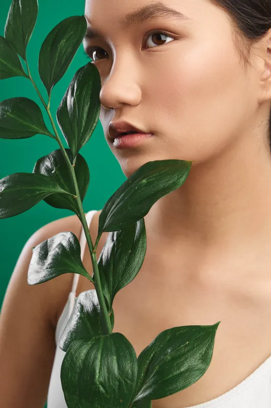 Asian woman gazes softly at the camera while holding a leafy plant near her face, a fresh green backdrop framing a calm, natural beauty in a studio portrait.