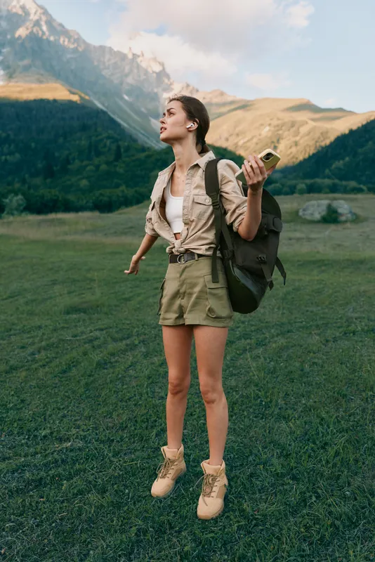 Young woman with a backpack stands in a green valley, looking up at the mountains while holding a phone, ready for adventure in a sunlit outdoor scene.