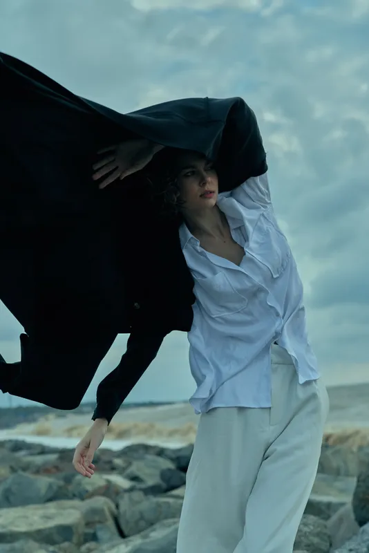 Woman on a windy beach pulls a dark coat over her head, white shirt and pale pants billowing, capturing motion, drama, and bold contrast in an outdoor coastal scene