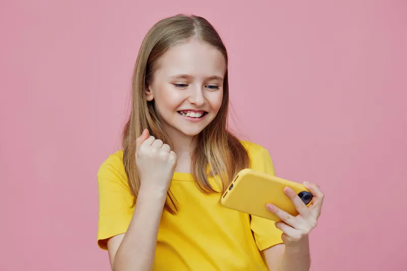 Happy girl in a yellow shirt celebrating with her smartphone against a pink background, expressing joy and excitement about a positive moment