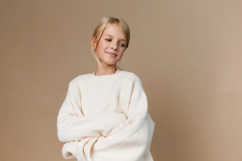 Authentic lifestyle portrait of a young girl in a soft cream sweater, capturing playful confidence and candid charm in a warm, minimalist studio setting.