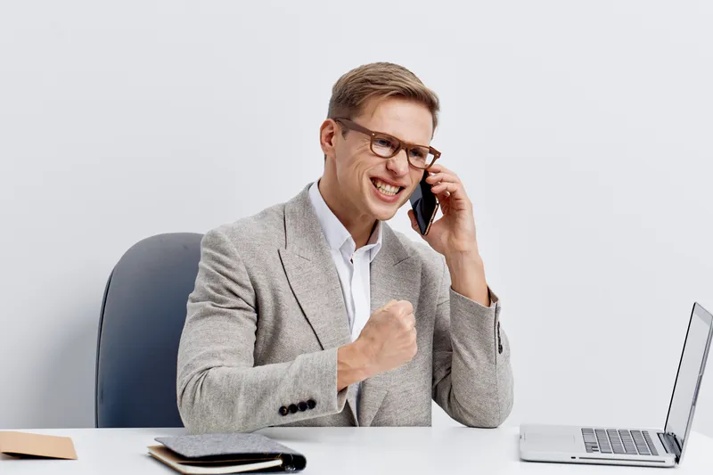 Professional man in glasses and suit smiling while talking on smartphone in modern office environment, sitting at desk with laptop and notepad, expressing success and confidence, business communicatio
