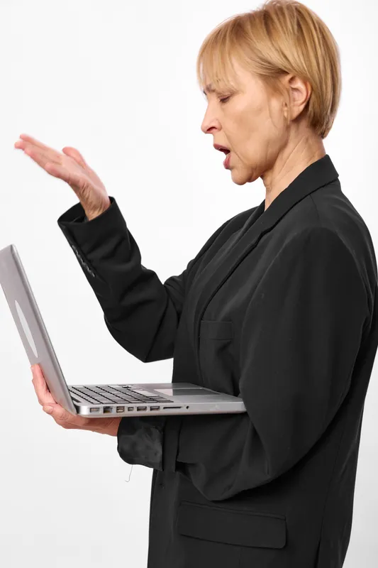 Elderly woman at laptop computer in office setting, working and communicating with expressive hand gesture, focused face, isolated on white background